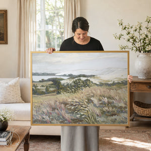 A framed landscape painting depicting a serene coastal scene. The artwork features a muted color palette of greens, browns, and blues, with a focus on grassy fields and distant hills. The frame is a warm, natural wood.
