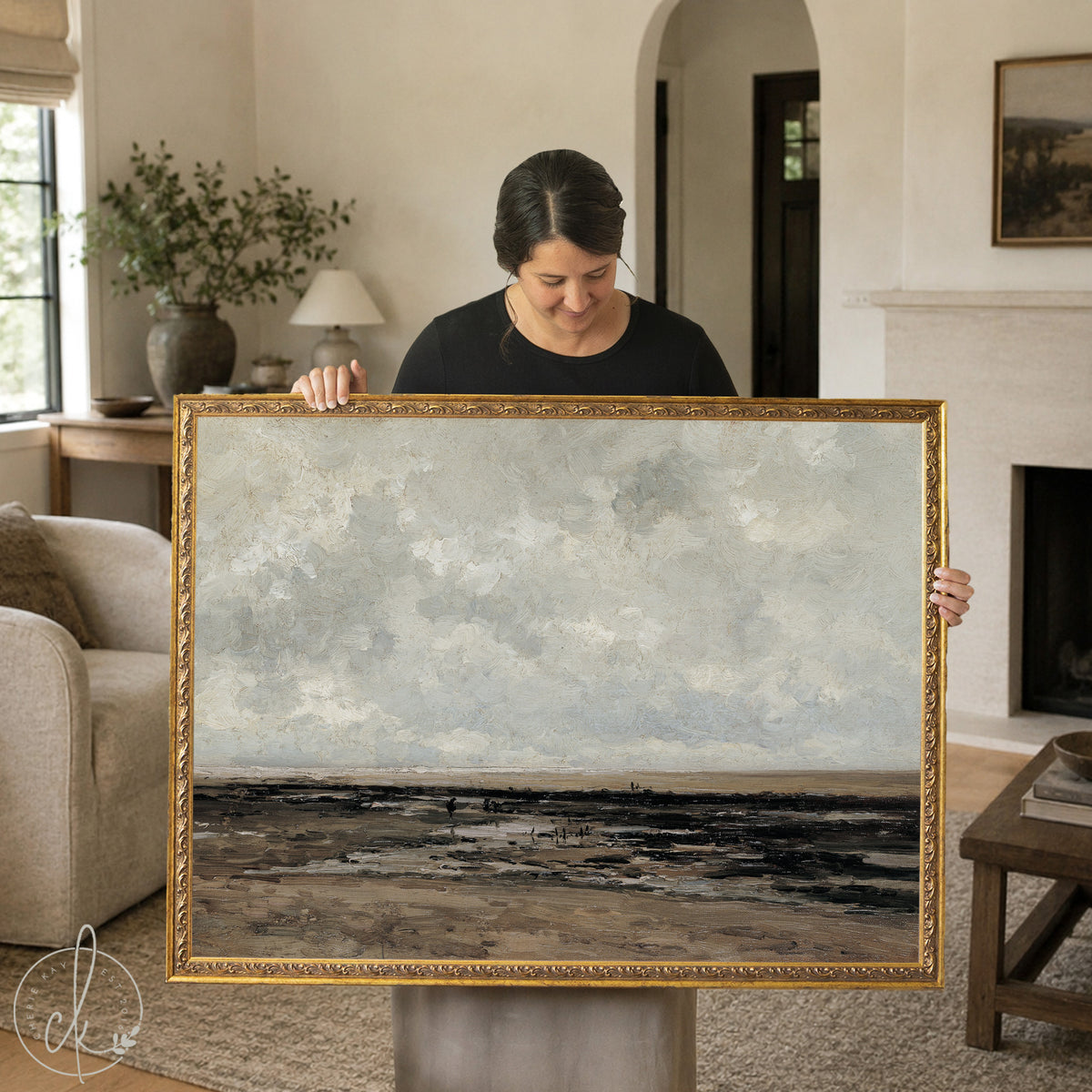 Woman holding a large vintage framed landscape painting featuring a moody coastal scene in earthy tones, ideal wall art for traditional or modern vintage home decor.