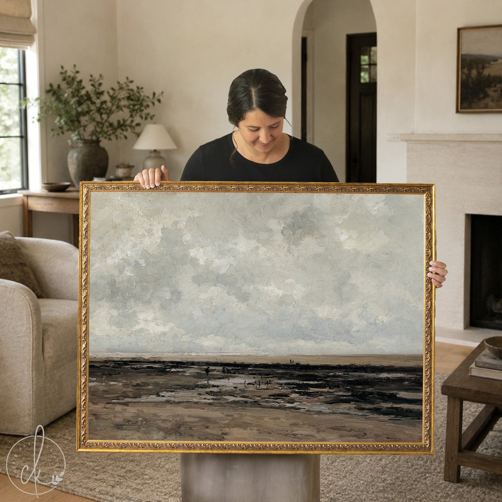 Woman holding a large vintage framed landscape painting featuring a moody coastal scene in earthy tones, ideal wall art for traditional or modern vintage home decor.