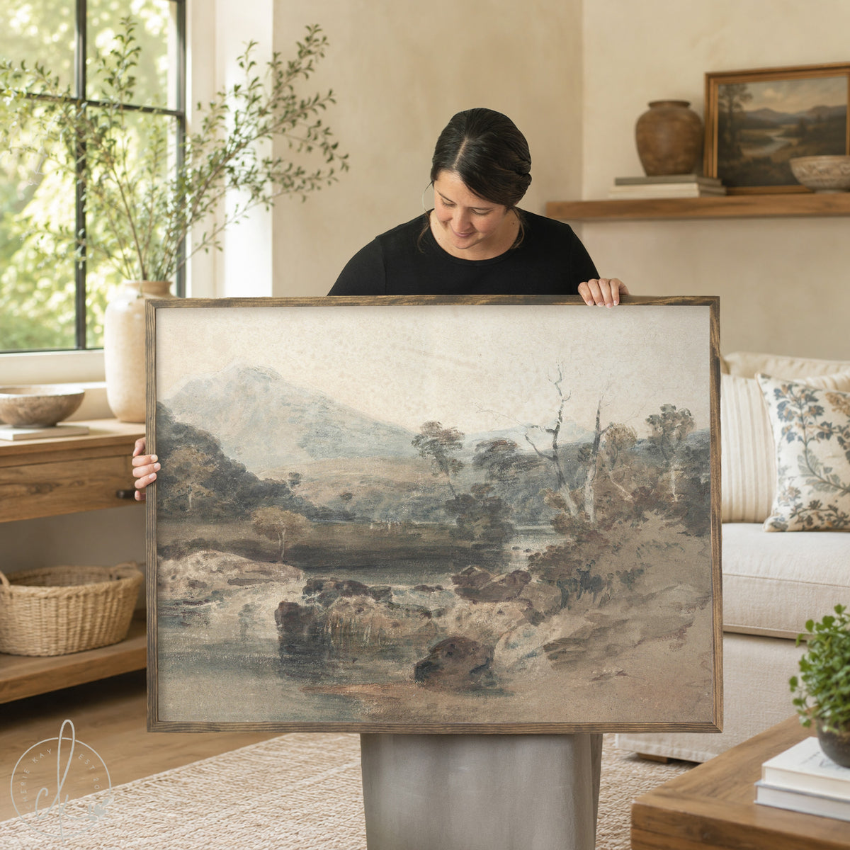 A woman holds up a large framed landscape painting in a bright, modern living room. The painting depicts a misty mountain and river scene in muted earth tones of gray, brown, and beige.