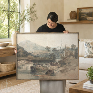 A woman holds up a large framed landscape painting in a bright, modern living room. The painting depicts a misty mountain and river scene in muted earth tones of gray, brown, and beige.