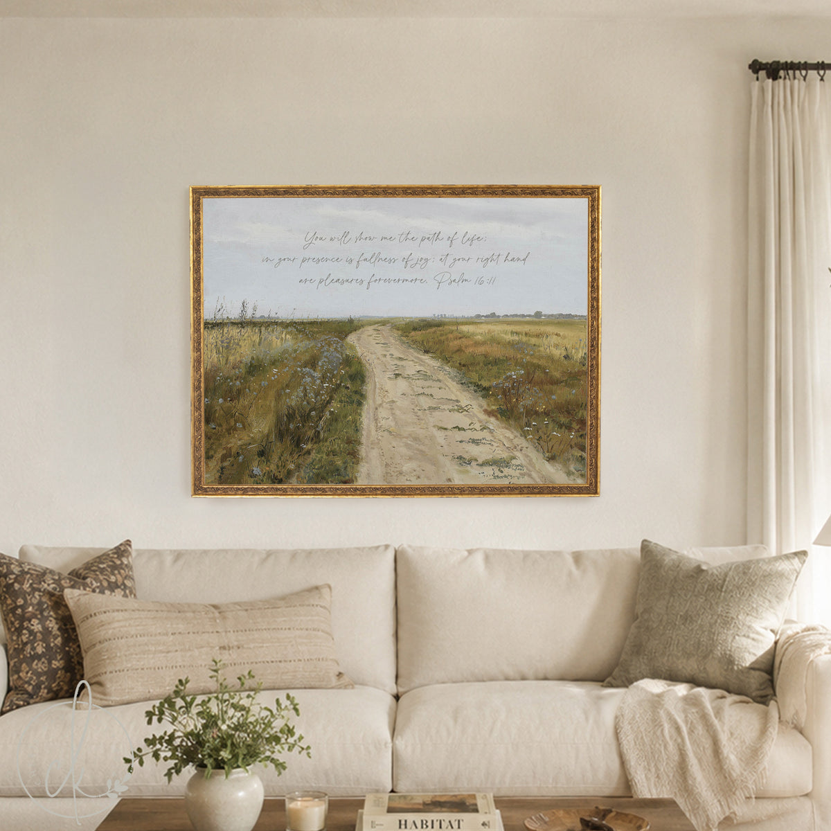 A framed art print featuring a scenic landscape with a dirt path through a field and a handwritten Bible verse, displayed on a beige wall above a cream-colored sofa in a living room setting.