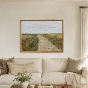A framed art print featuring a scenic landscape with a dirt path through a field and a handwritten Bible verse, displayed on a beige wall above a cream-colored sofa in a living room setting.