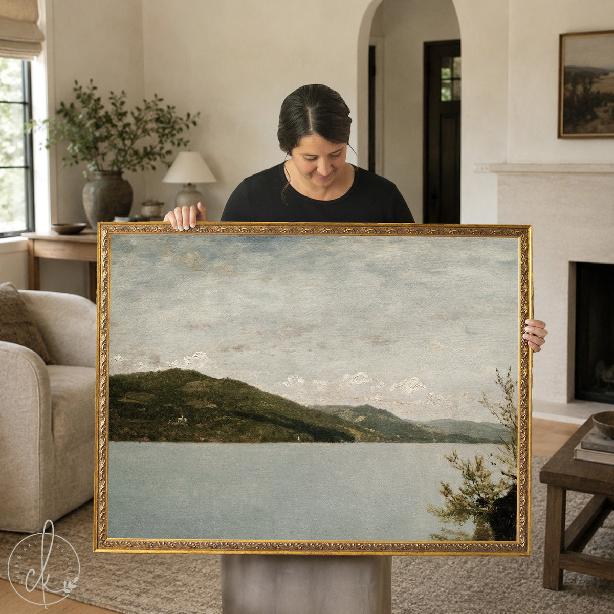 A woman stands in front of a house, holding a large vintage framed oil painting of a lake and mountain. The artwork features a calm, muted blue lake bordered by a tree-covered hillside, with a range of distant mountains fading into the soft, cloudy sky. The frame has a classic, dark finish that complements the natural tones of the artwork.