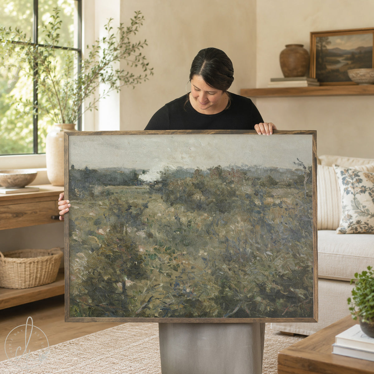 Large landscape wall art featuring abstract countryside scene with rolling hills and muted earth tones, displayed by woman in modern living room setting with neutral decor and natural wood frame
