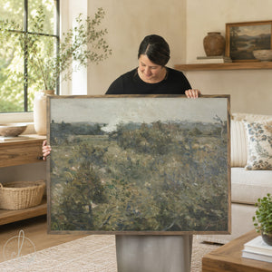 Large landscape wall art featuring abstract countryside scene with rolling hills and muted earth tones, displayed by woman in modern living room setting with neutral decor and natural wood frame