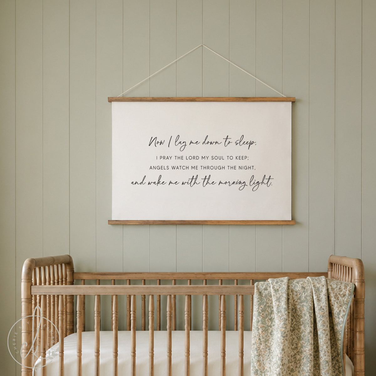 A rectangular wall hanging featuring a cream-colored fabric banner suspended by two light wood rails and a thin string. The banner displays the black printed text: Now I lay me down to sleep. I PRAY THE LORD MY SOUL TO KEEP; ANGELS WATCH ME THROUGH THE NIGHT, and wake me with the morning light.