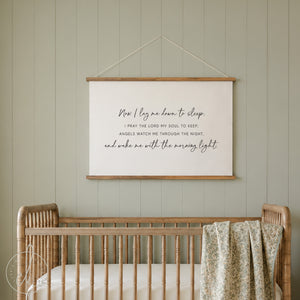 A rectangular wall hanging featuring a cream-colored fabric banner suspended by two light wood rails and a thin string. The banner displays the black printed text: Now I lay me down to sleep. I PRAY THE LORD MY SOUL TO KEEP; ANGELS WATCH ME THROUGH THE NIGHT, and wake me with the morning light.