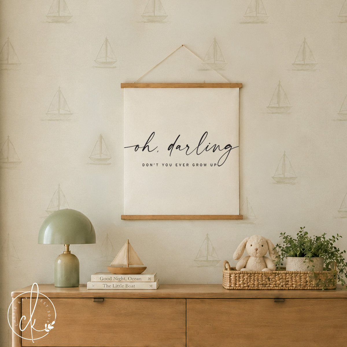 A rectangular wall hanging featuring a cream-colored fabric canvas held by light wood rails at the top and bottom. The text "oh, darling" is printed in a black script font above the text "DON'T YOU EVER GROW UP" in black.