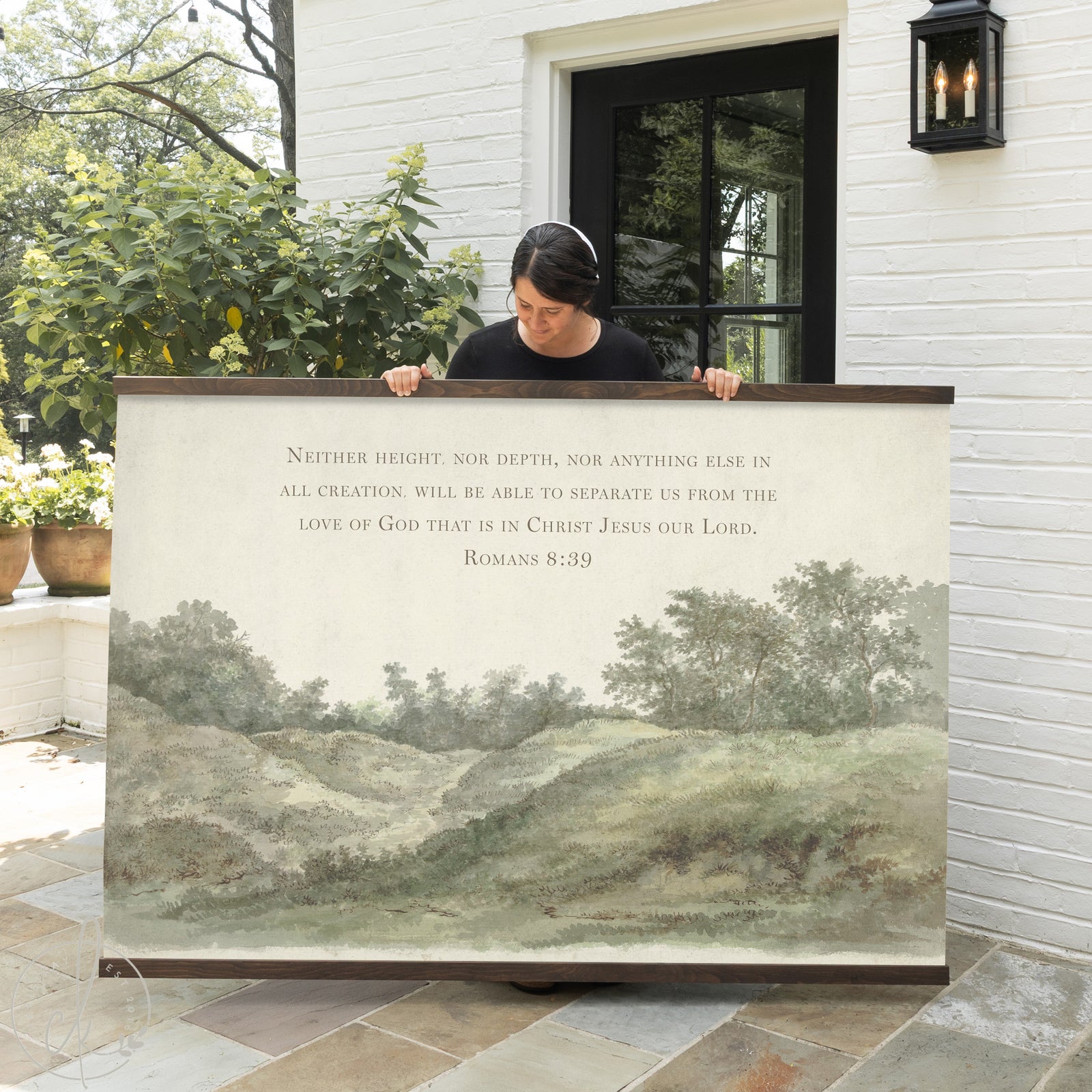 A person holding wall art of a countryside landscape with a verse from Romans 8:39. The background features a sofa and a potted plant.