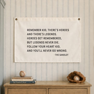 A white rectangular fabric wall hanging featuring black printed text that reads: REMEMBER KID, THERE'S HEROES AND THERE'S LEGENDS. HEROES GET REMEMBERED, BUT LEGENDS NEVER DIE. FOLLOW YOUR HEART KID, AND YOU'LL NEVER GO WRONG. - THE SANDLOT.