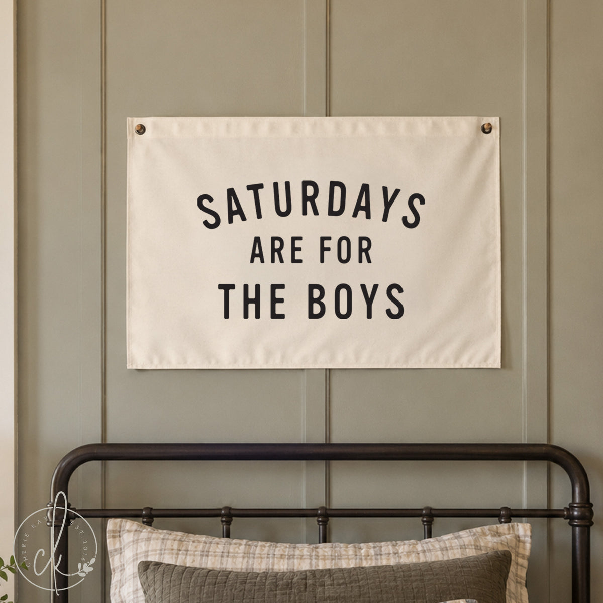 A rectangular white fabric wall banner featuring the black text SATURDAYS ARE FOR THE BOYS in a bold sans-serif font. The banner is secured with metal grommets in the top corners and over a little boys bed. 