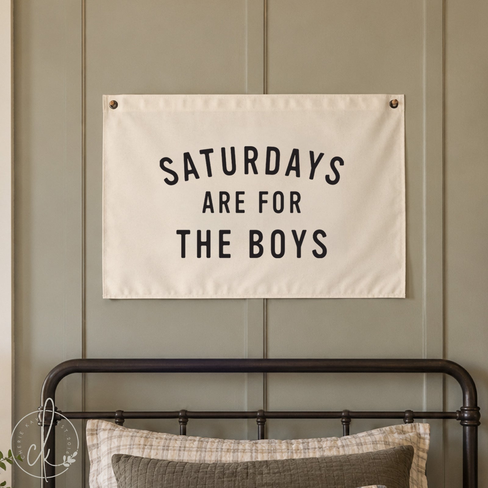 A rectangular white fabric wall banner featuring the black text SATURDAYS ARE FOR THE BOYS in a bold sans-serif font. The banner is secured with metal grommets in the top corners and over a little boys bed. 