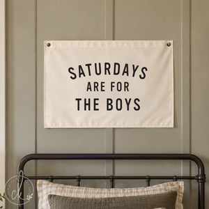 A rectangular white fabric wall banner featuring the black text SATURDAYS ARE FOR THE BOYS in a bold sans-serif font. The banner is secured with metal grommets in the top corners and over a little boys bed. 