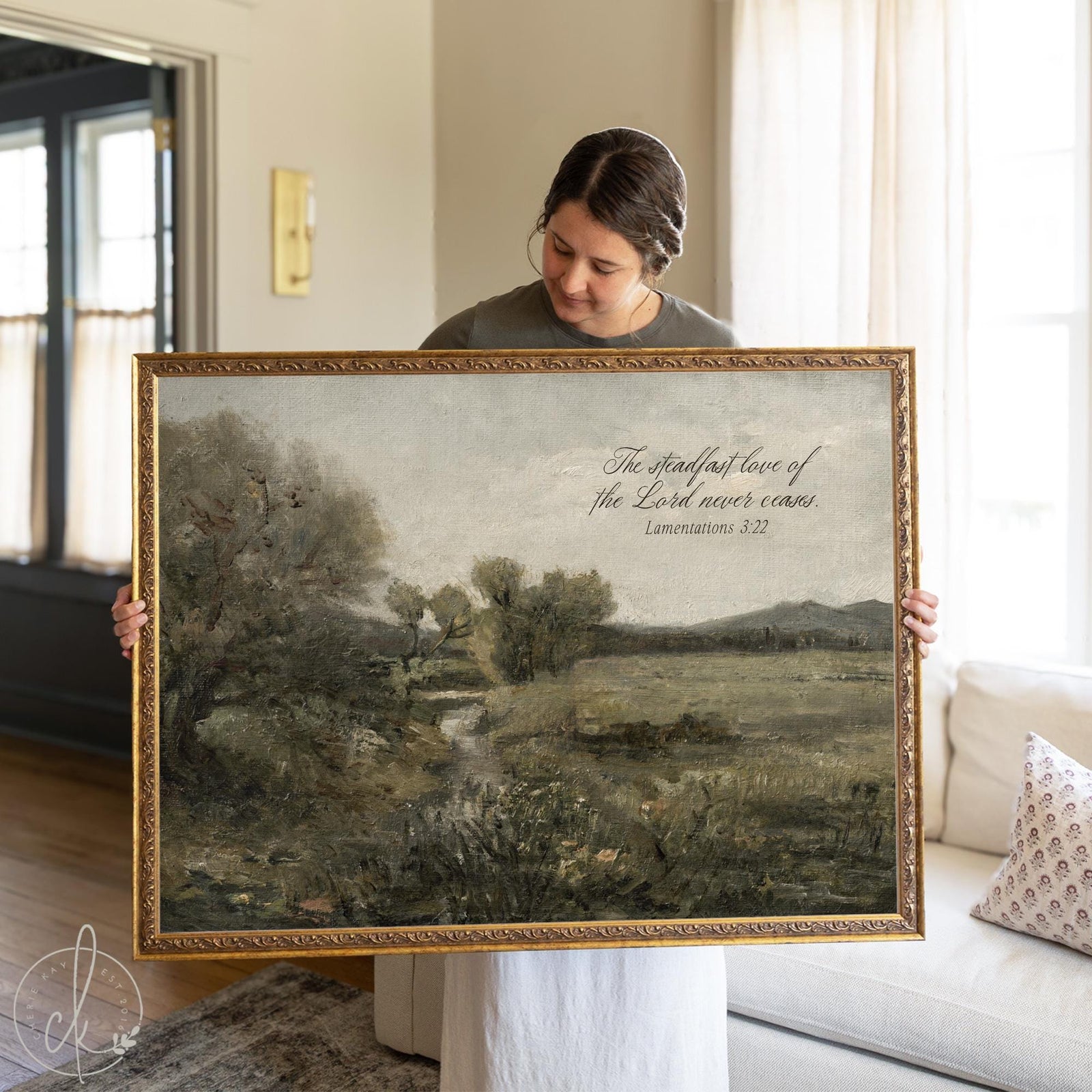Large wall art with scripture featuring Lamentations 3:22 &#39;The steadfast love of the Lord never ceases&#39; displayed over vintage landscape painting in ornate gold frame, shown by woman in bright living room