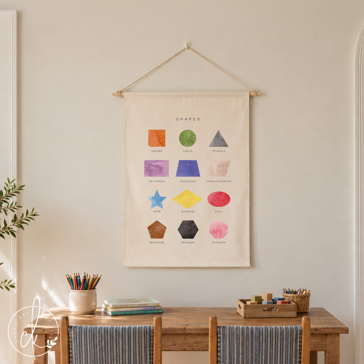 A cream-colored fabric wall hanging featuring the text SHAPES and twelve watercolor illustrations of geometric forms including a square, circle, triangle, rectangle, trapezoid, parallelogram, star, diamond, oval, pentagon, hexagon, and octagon.