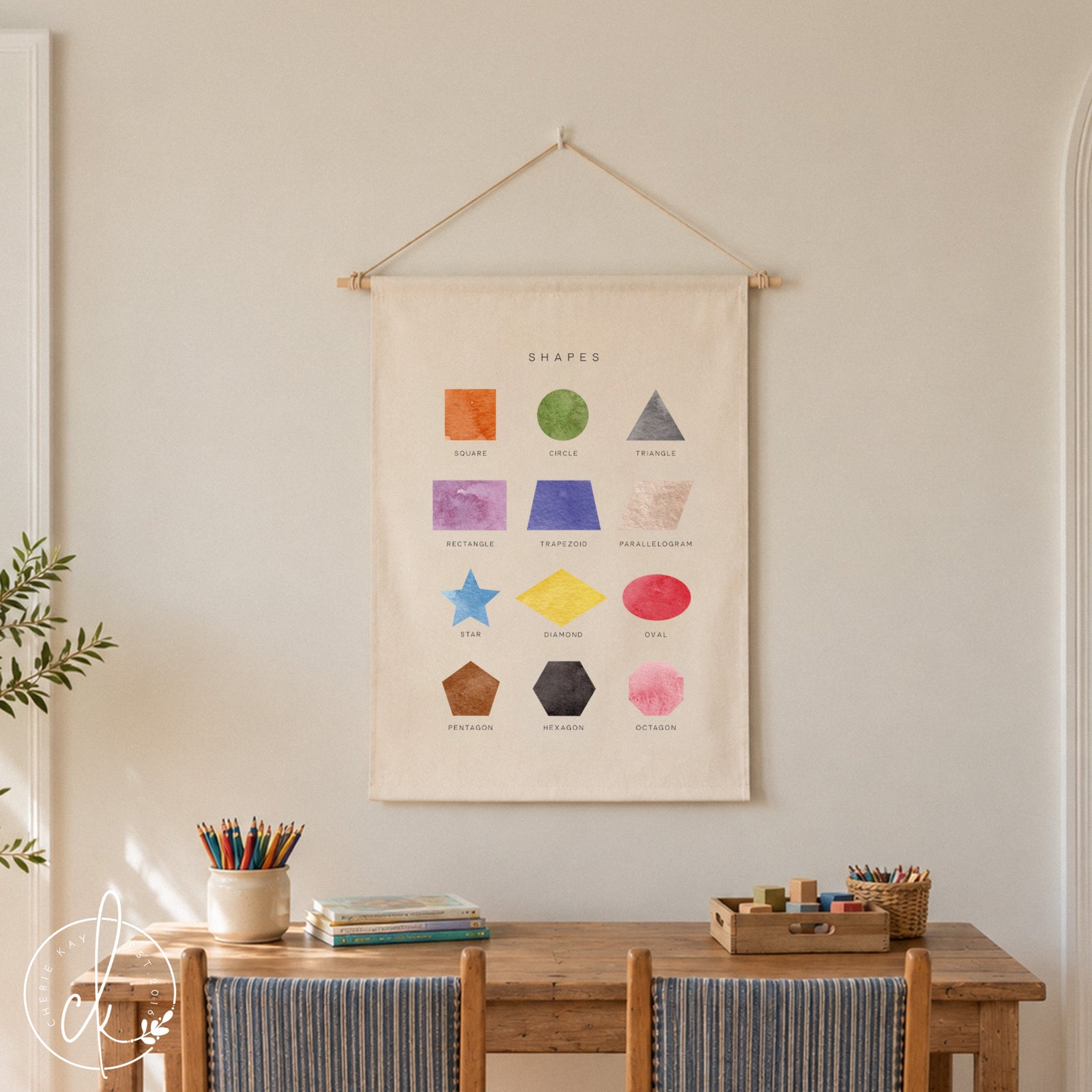 A cream-colored fabric wall hanging featuring the text SHAPES and twelve watercolor illustrations of geometric forms including a square, circle, triangle, rectangle, trapezoid, parallelogram, star, diamond, oval, pentagon, hexagon, and octagon.