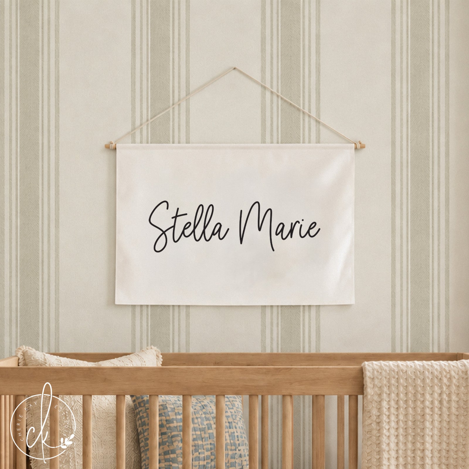 A rectangular off-white fabric wall hanging banner featuring the black cursive text Stella Marie. The banner is suspended from a thin wooden rod with a white string attached to the ends for hanging on a wall above a wooden crib.