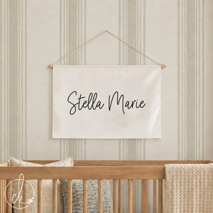 A rectangular off-white fabric wall hanging banner featuring the black cursive text Stella Marie. The banner is suspended from a thin wooden rod with a white string attached to the ends for hanging on a wall above a wooden crib.