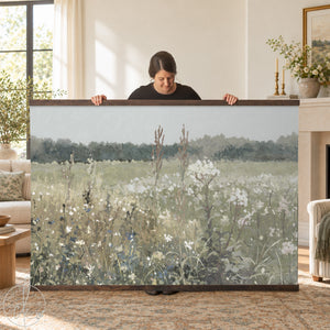 A large rectangular wall art print featuring a landscape painting of a field with wildflowers and tall grasses in muted green, beige, and white tones, framed with dark brown wooden rails at the top and bottom edges.