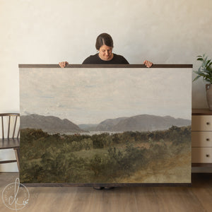 A large rectangular landscape wall art print featuring a painting of rolling hills, a distant body of water, and lush green trees under a cloudy sky, secured between two dark brown wooden magnetic hanging rails.