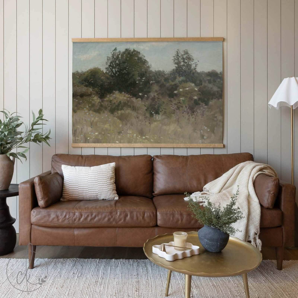 A cozy living room with a brown leather sofa, a golden coffee table, and a potted plant, featuring a large painting of trees and foliage on the wall.