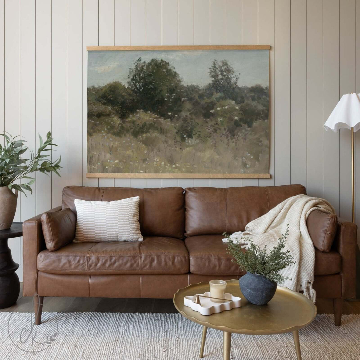 A cozy living room with a brown leather sofa, a golden coffee table, and a potted plant, featuring a large painting of trees and foliage on the wall.