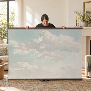 A large rectangular wall hanging featuring a printed image of white and light gray clouds against a pale blue sky. The print is secured between two horizontal dark brown wooden bars at the top and bottom edges.