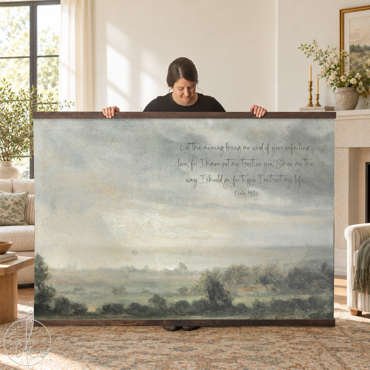 A person holding wall art of a serene landscape with a verse from Psalm 143:8. The background features a sofa and a potted plant.