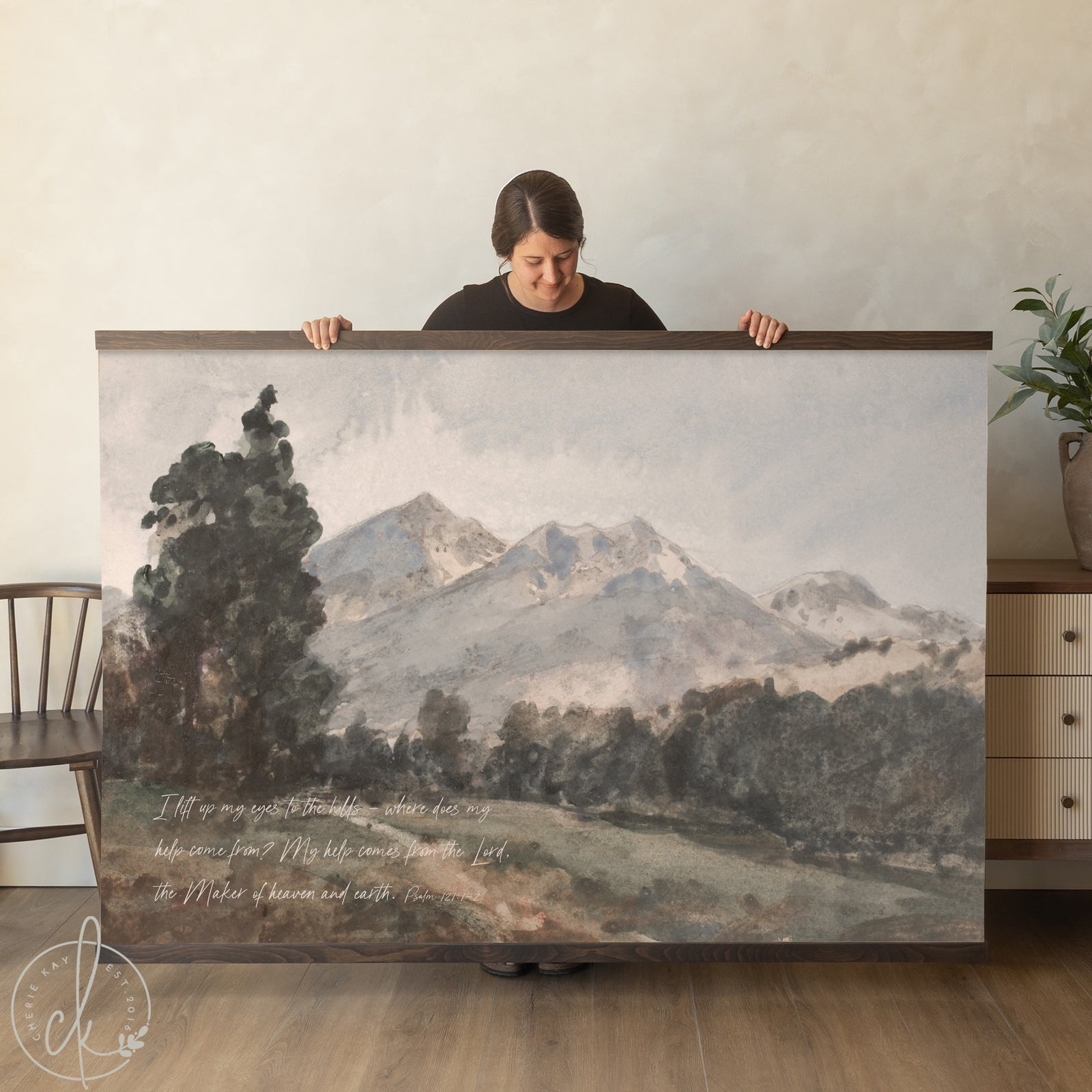 A person holding a wall art piece of a mountain landscape with a verse from Psalm 121:1-2. The background features a neutral interior with a sofa and plant.