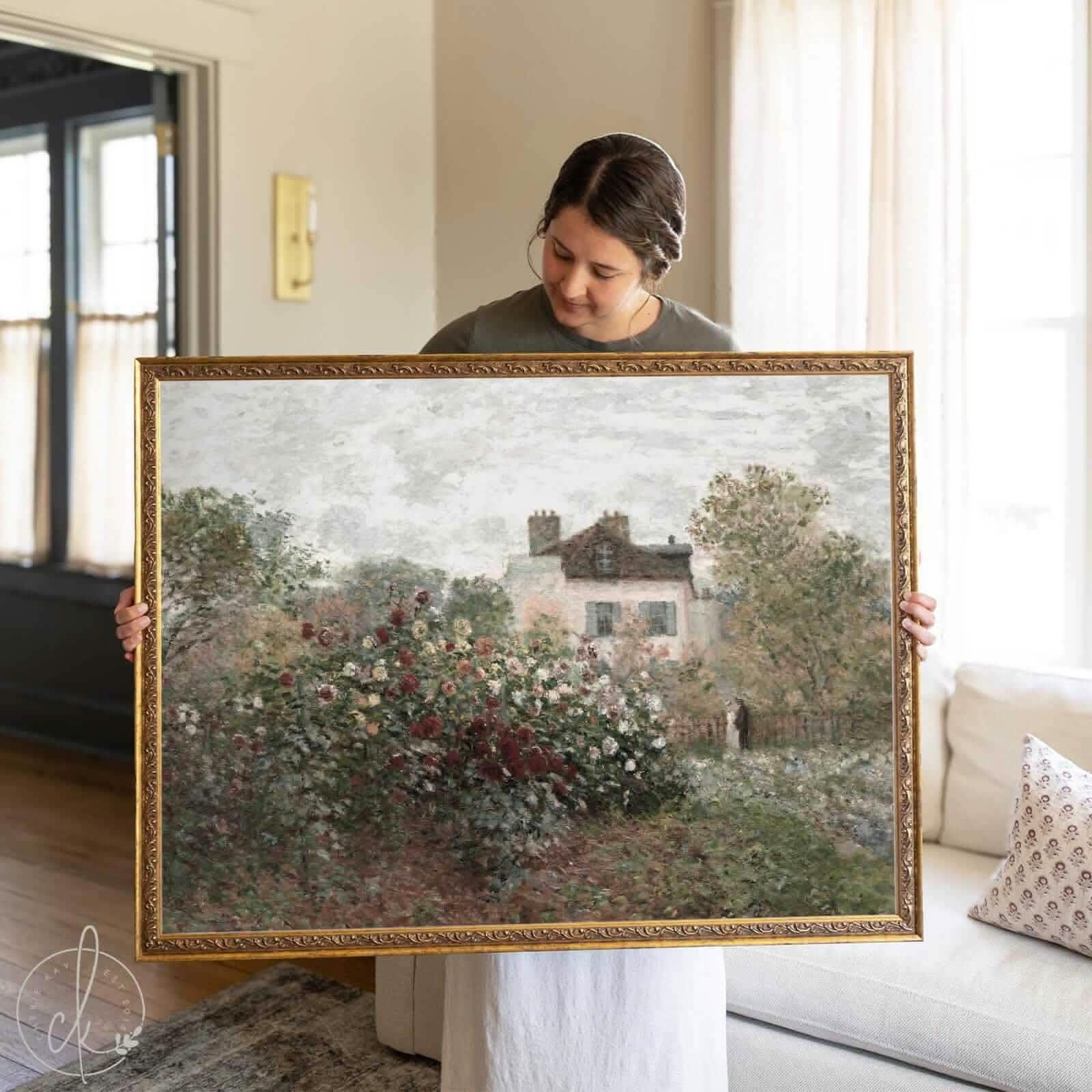 Woman holding a large framed vintage-style landscape art print featuring a blooming garden and French country house, inspired by Claude Monet impressionist style.