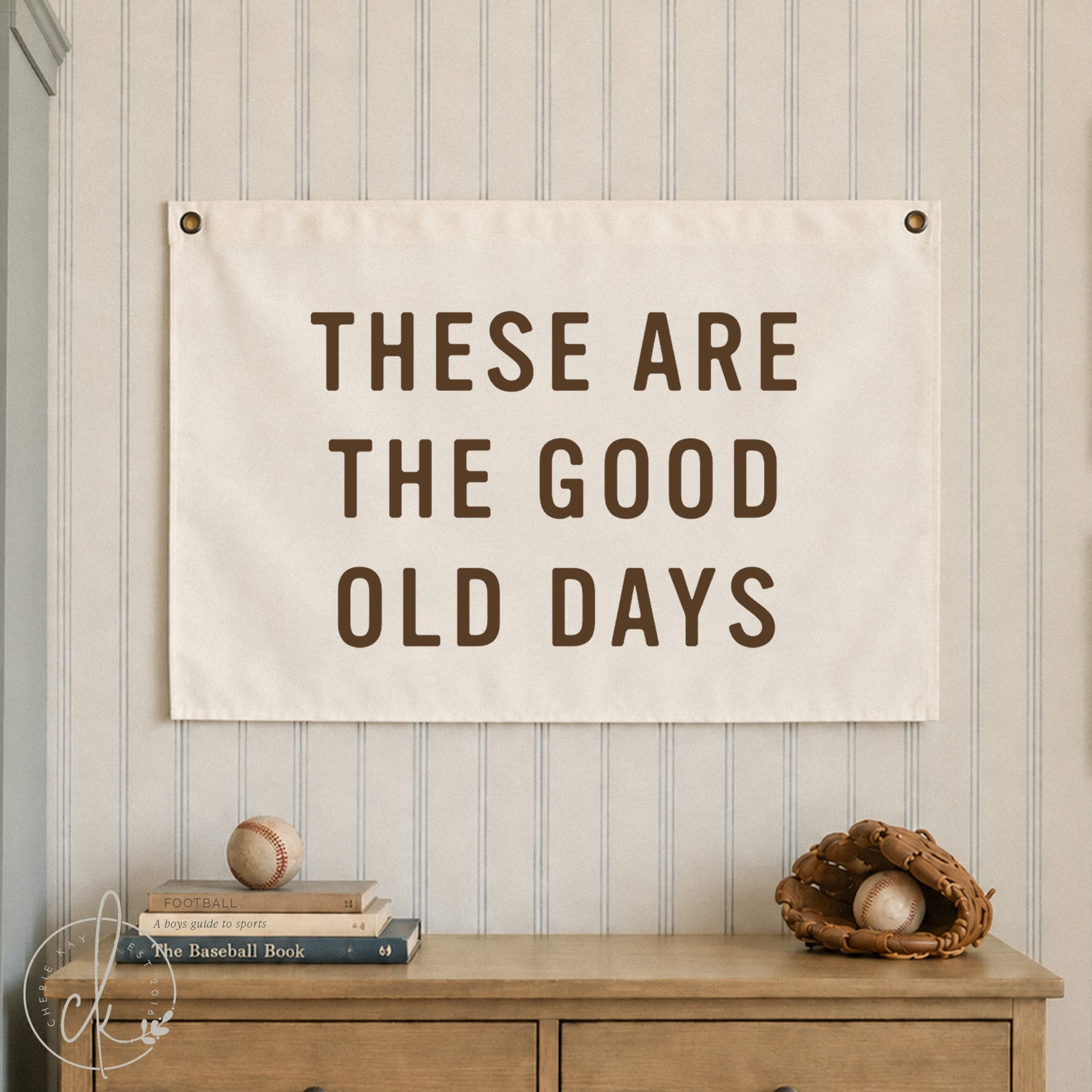 A canvas banner with the text 'THESE ARE THE GOOD OLD DAYS' in brown lettering, hung on a beige and white striped wall above a wooden dresser with baseball-themed props.