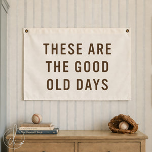 A canvas banner with the text 'THESE ARE THE GOOD OLD DAYS' in brown lettering, hung on a beige and white striped wall above a wooden dresser with baseball-themed props.