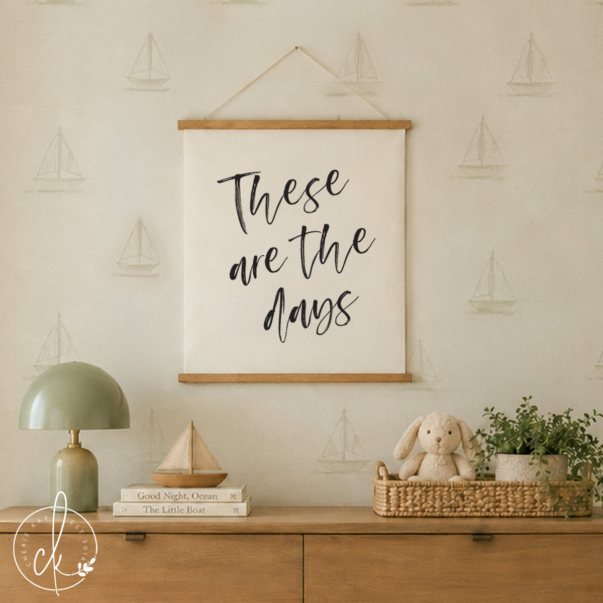 A white wooden crib with a white sheet, two beige pillows, and a cream-colored waffle-knit blanket. Above the crib hangs a white canvas wall hanging with wooden rails and the black text "These are the days" in a script font.