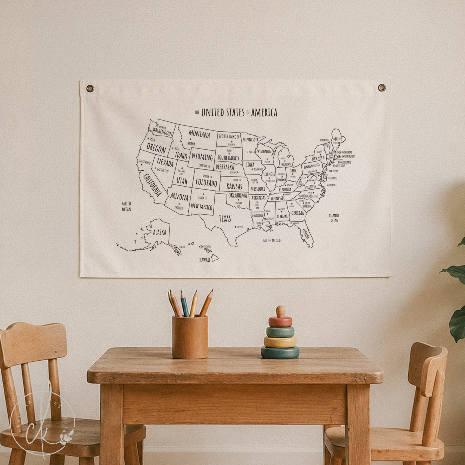A fabric wall tapestry featuring a black line-art map of the United States hanging on a beige wall above a wooden children's table and chairs.