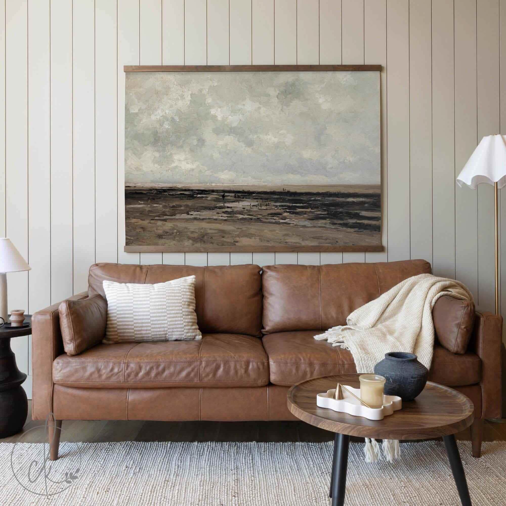 Neutral-toned landscape tapestry wall art featuring a vintage coastal scene with textured brushstrokes, framed with a wooden hanger, displayed above a brown leather sofa in a cozy living room setting.