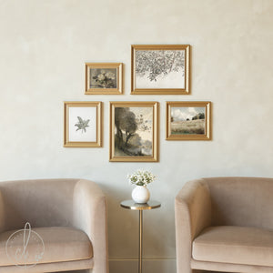 Five gold-framed vintage artworks displayed in a curated gallery wall arrangement above neutral beige armchairs, featuring botanical illustrations, pastoral landscapes, a serene river scene, and a delicate floral still life, creating a cohesive collection of nature-inspired art.