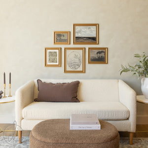 A neutral living room featuring a cream textured sofa with a brown grid-patterned pillow, positioned below a gallery wall of five gold-framed landscape and nature artworks arranged asymmetrically, with a round beige ottoman displaying books in the foreground.