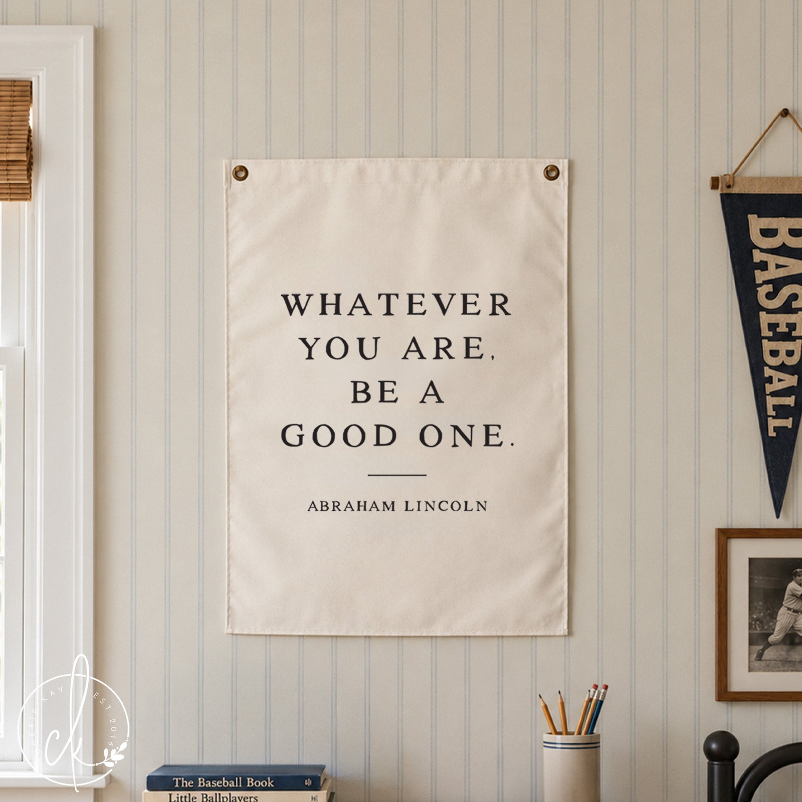 A white rectangular wall hanging featuring the black text: WHATEVER YOU ARE, BE A GOOD ONE. ABRAHAM LINCOLN. The sign is mounted with two metal grommets at the top corners, displaying a clear, bold, serif typeface on a clean white background.