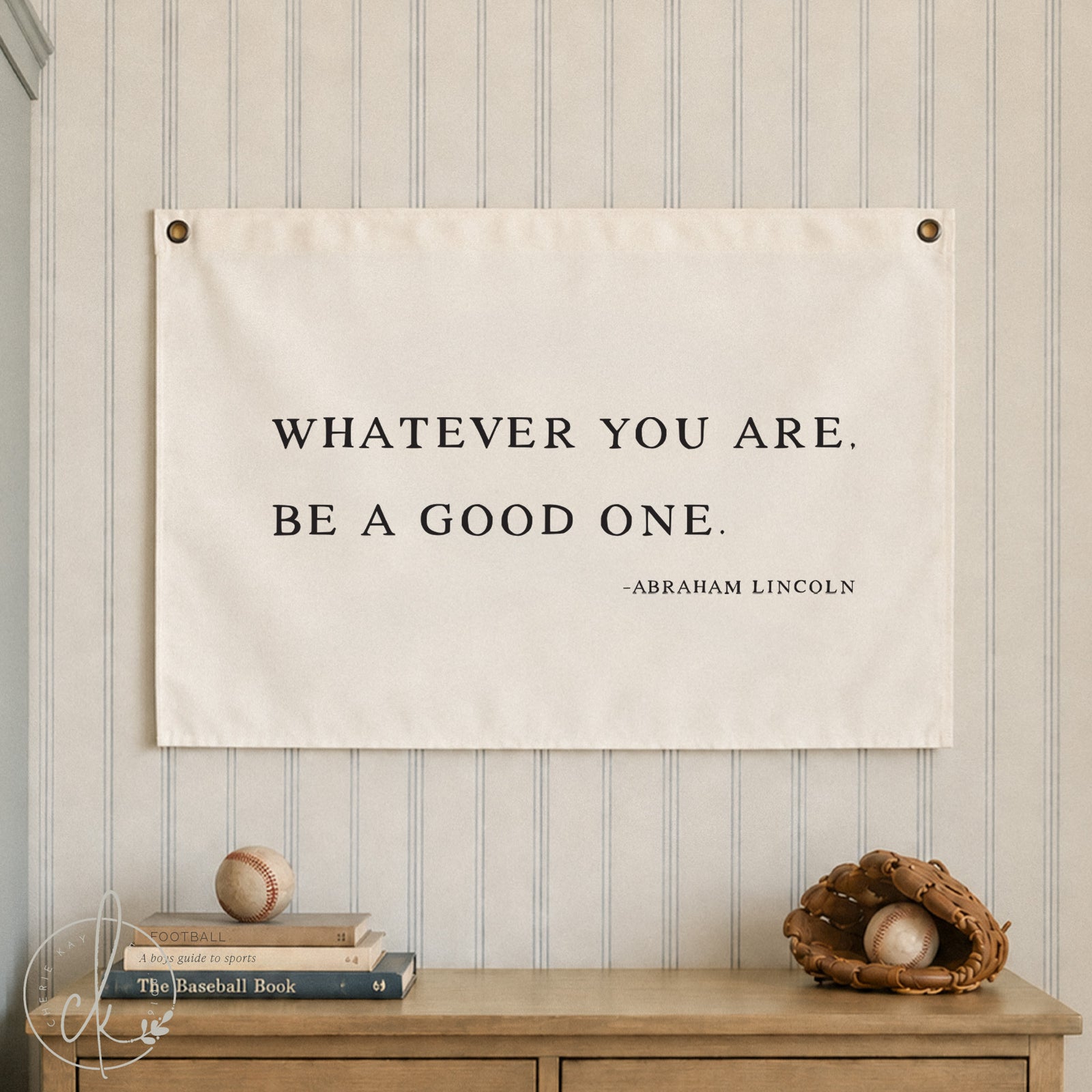 A rectangular white wall banner featuring the black printed text: WHATEVER YOU ARE. BE A GOOD ONE. -ABRAHAM LINCOLN. The banner is secured with metal grommets in the top corners and displays a clear, bold, serif typeface on a fabric surface.