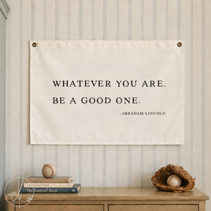 A rectangular white wall banner featuring the black printed text: WHATEVER YOU ARE. BE A GOOD ONE. -ABRAHAM LINCOLN. The banner is secured with metal grommets in the top corners and displays a clear, bold, serif typeface on a fabric surface.