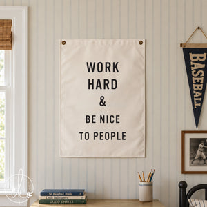 A white rectangular wall hanging featuring the black printed text WORK HARD & BE NICE TO PEOPLE. The sign is secured with two metal grommets at the top corners and displays a clean, bold sans-serif font against a plain white background.