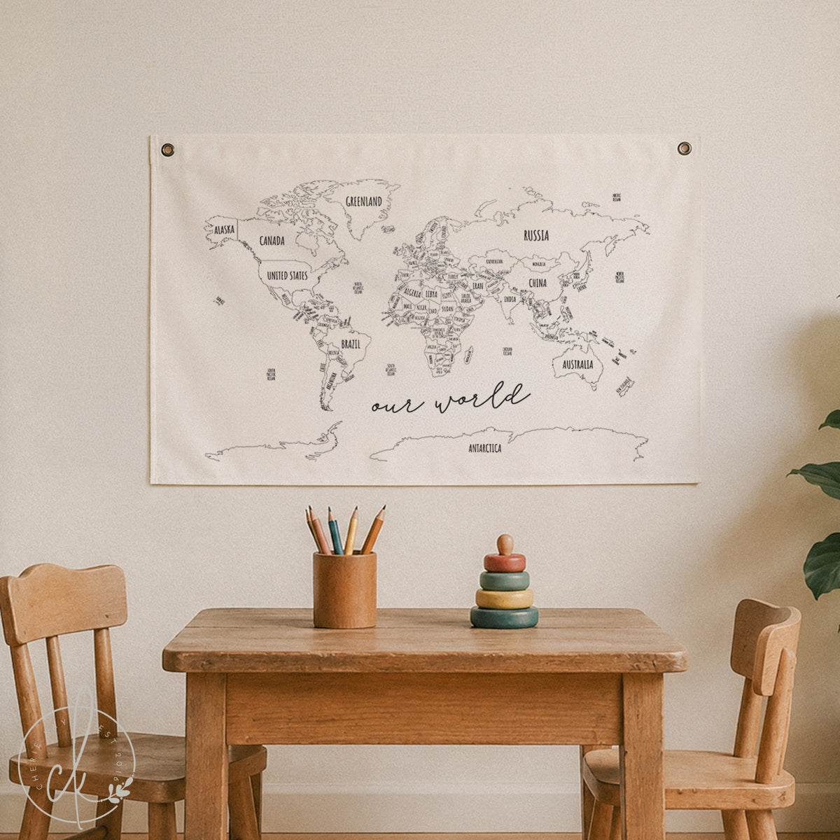 A large cream-colored fabric world map wall hanging with black line drawings and country names, displayed above a wooden table in a child's room setting with two wooden chairs.
