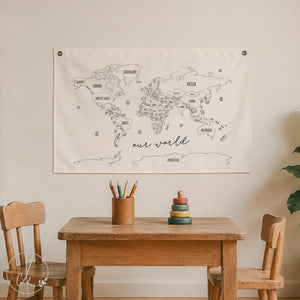 A large cream-colored fabric world map wall hanging with black line drawings and country names, displayed above a wooden table in a child's room setting with two wooden chairs.