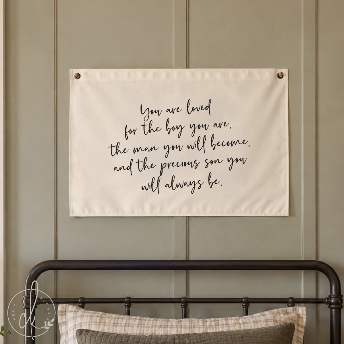 A rectangular off-white fabric wall hanging featuring black cursive text that reads: You are loved for the boy you are, the man you will become, and the precious son you will always be. The banner has metal grommets at the top corners.