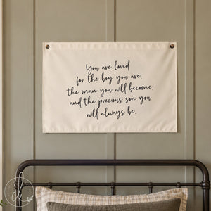 A rectangular off-white fabric wall hanging featuring black cursive text that reads: You are loved for the boy you are, the man you will become, and the precious son you will always be. The banner has metal grommets at the top corners.