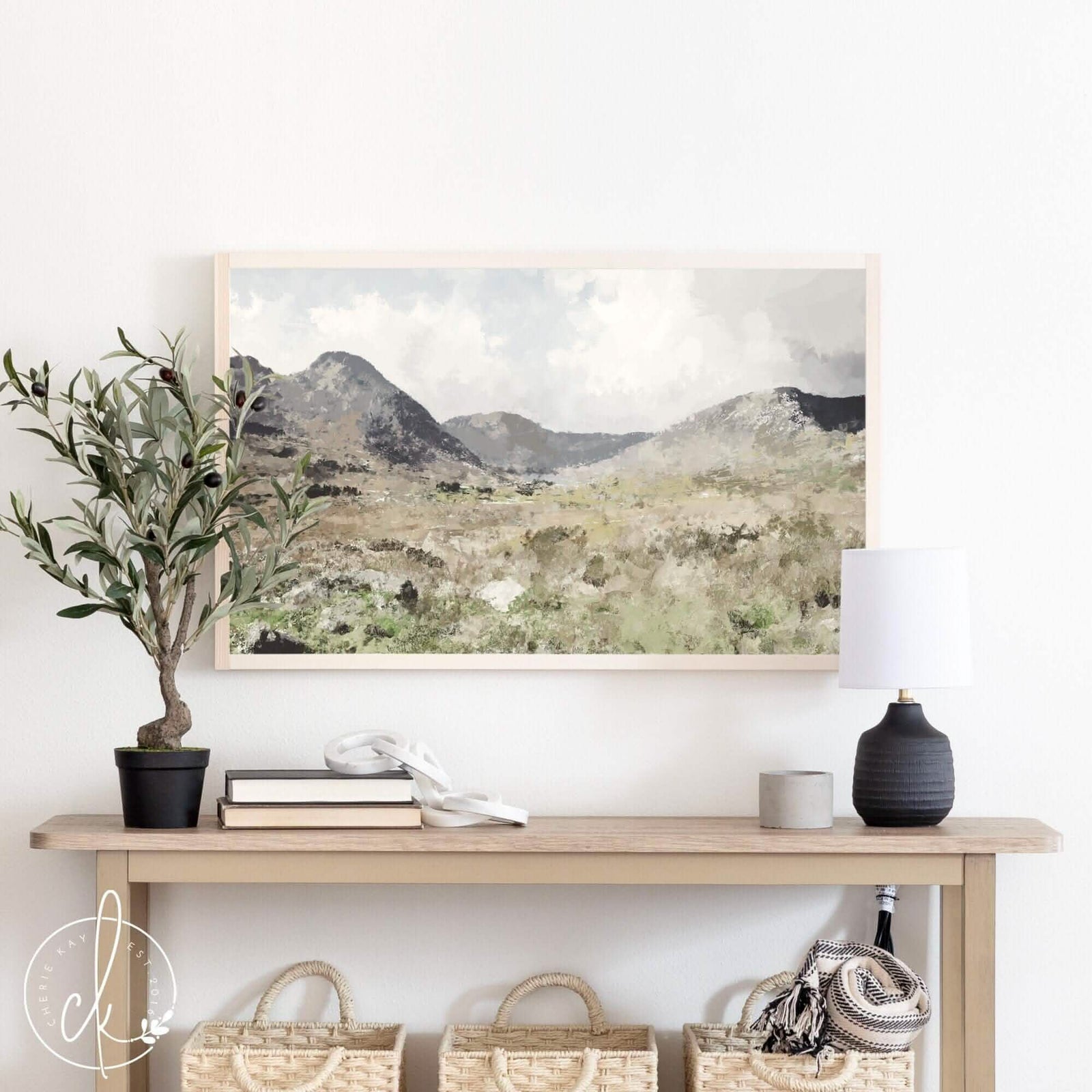 This is a painting of mountain wall art. The landscape has a meadow and mountains in the distance. The painting is framed in a pine frame and is displayed above a table with modern decor.