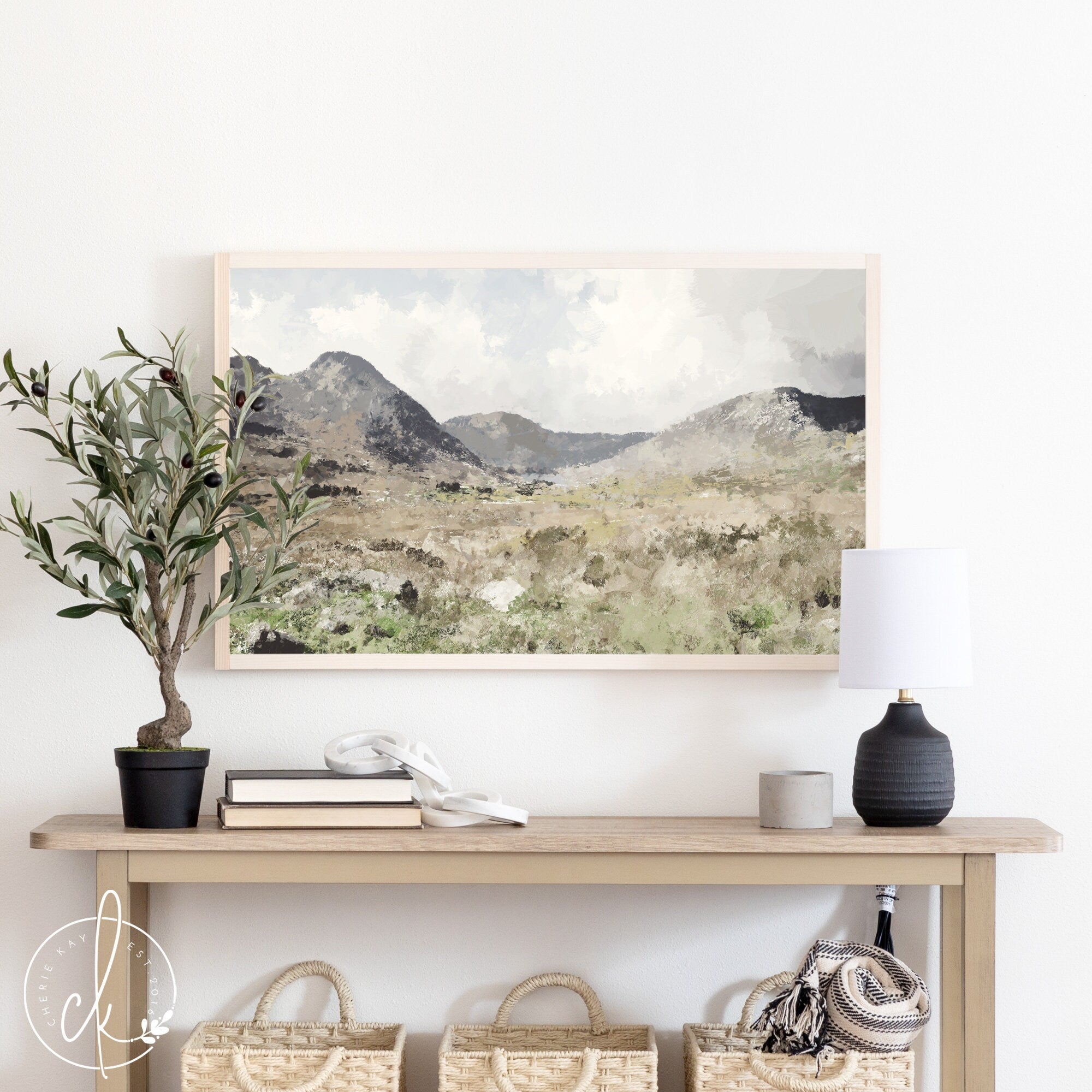 This is a painting of mountain wall art. The landscape has a meadow and mountains in the distance. The painting is framed in a pine frame and is displayed above a table with modern decor.