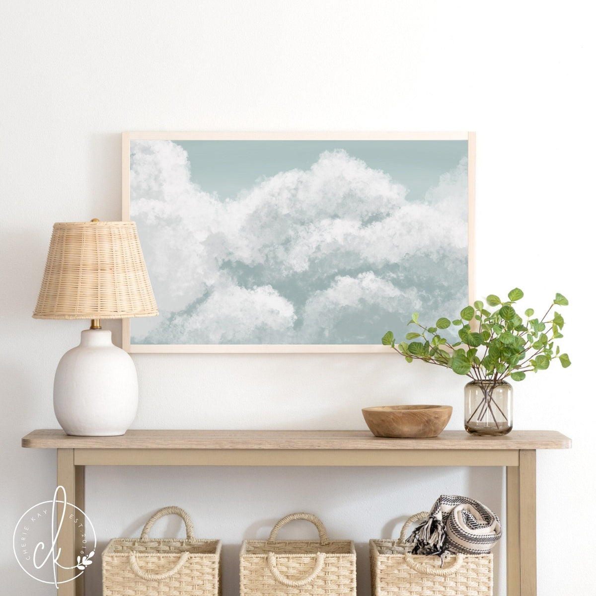A painting of blue sky and clouds on a sunny day. This painting is framed with a natural pine frame, and is displayed above a table with a lamp and other home decor.