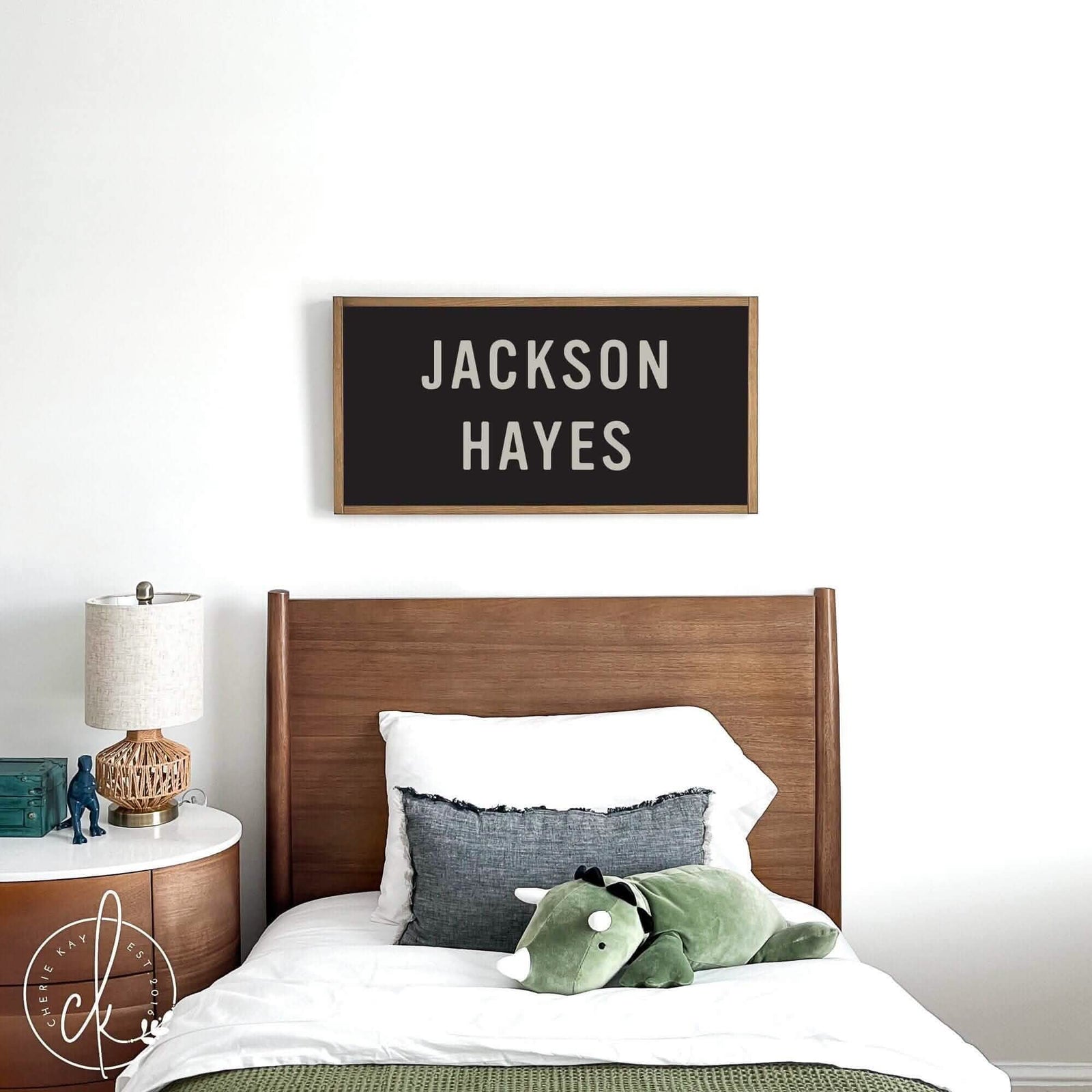 A wooden bed with white and gray bedding, a green stuffed animal, and a personalized name sign "JACKSON HAYES" above the headboard.